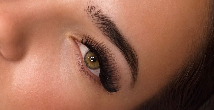 Close-up of a woman's eye showcasing long, dark, curled false eyelashes, with her eyebrow and skin visible, resting on a pink fluffy surface.